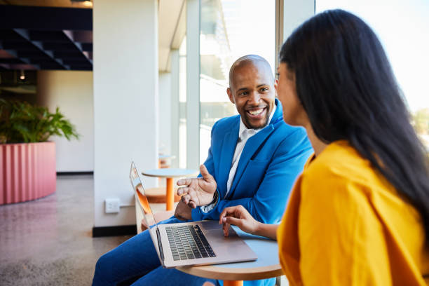 Smiling young businessman discussing work on a laptop with a female coworker at a table in the hallway of a modern office